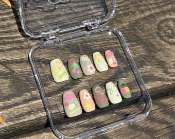 May include: A clear plastic case with a hinged lid containing ten false fingernails with floral designs. The designs include flowers, hearts, and the words "love" and "summer".