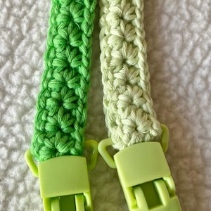 May include: Two green crocheted straps with lime green plastic clips. The straps are made with a simple stitch pattern and are attached to the clips with a loop.