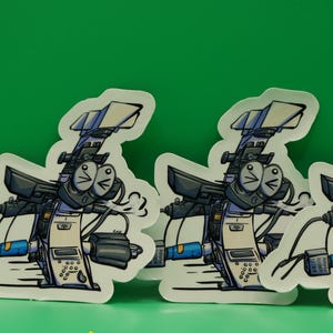 May include: Three vinyl stickers featuring a cartoon camera character with a smiling face, black and white details, and blue accents. The stickers are on a green background with the words "VINYL STICKER" in yellow.