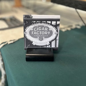 May include: Black and white photo of a sign that reads "Cigar Factory Charleston" with a logo of a "C" and "F" intertwined. The sign is on a black stand and the background is blurred.