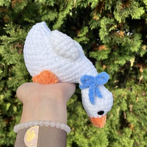 May include: A handmade white crochet goose with orange feet and beak, and a blue bow. The goose is held up against a backdrop of green foliage. The craft item is soft and plush.