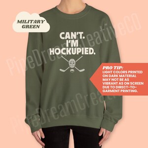 May include: A dark green sweatshirt with the text "CAN'T. I'M HOCKUPIED." in white. A hockey stick and mask graphic is below the text. The text "PRO TIP: LIGHT COLORS PRINTED ON DARK MATERIAL MAY NOT BE AS VIBRANT AS ON SCREEN DUE TO DIRECT-TO-GARMENT PRINTING." is in the bottom right corner.