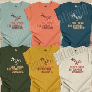 May include: A collection of six t-shirts in various colors, including teal, orange, navy, olive green, yellow, and cream. Each shirt features the text "I DON'T CRASH. I DO 'SURPRISE DISMOUNTS.'" along with a graphic of a person and a bicycle.