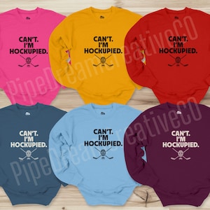 May include: Six long-sleeved sweatshirts in different colors with the text "CAN'T. I'M HOCKUPIED." and a hockey stick and mask graphic.