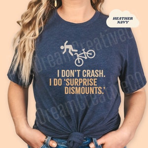 May include: Heather navy blue t-shirt with a bicycle graphic and the text "I DON'T CRASH. I DO 'SURPRISE DISMOUNTS.'" The shirt features a crew neck and short sleeves. The graphic shows a person falling off a bicycle.