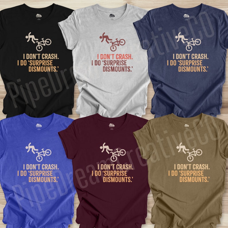 May include: A collection of t-shirts in various colors, including black, gray, blue, and burgundy. Each shirt features a graphic of a person and a bicycle, along with the text "I DON'T CRASH. I DO 'SURPRISE DISMOUNTS.'"