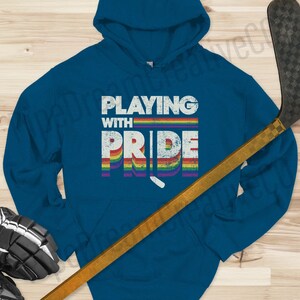May include: A blue hoodie with a rainbow design and the text "Playing with Pride". A hockey stick is in the foreground.