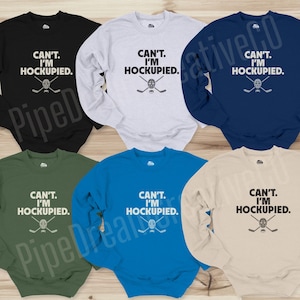 May include: Six different color long-sleeve sweatshirts with a hockey stick and puck graphic and the text "CAN'T. I'M HOCKUPIED."