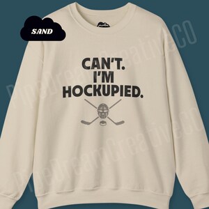 May include: A cream-colored sweatshirt with the text "CAN'T. I'M HOCKUPIED." in black. A hockey mask and crossed hockey sticks are below the text.