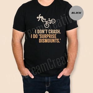 May include: Black t-shirt with a bicycle rider graphic and the text "I DON'T CRASH. I DO 'SURPRISE DISMOUNTS.'" The shirt is a crew neck style.