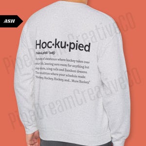 May include: A gray long-sleeve sweatshirt with the text "Hoc-ku-pied" defined as "a state of existence where hockey takes over your life, leaving zero room for anything but slap shots, icing calls and Zamboni dreams. The condition where your schedule reads: "Hockey, Hockey, Hockey, and... More Hockey.""