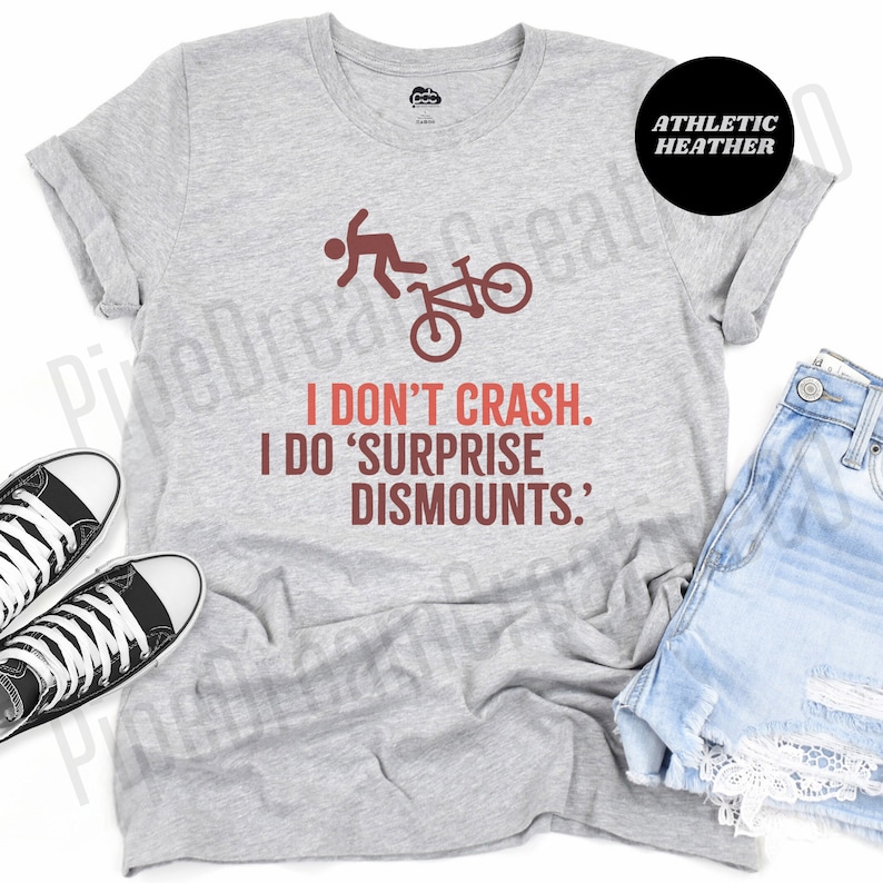 May include: Athletic heather gray t-shirt with the text "I don't crash. I do 'surprise dismounts'" in red and brown. The shirt features a graphic of a person flying off a bicycle. The shirt is paired with denim shorts and black and white sneakers.