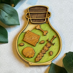 May include: Wooden potion bottle with a green interior, featuring a treasure chest, skull, bone, coins, and a sword. The bottle has a wooden lid and a light brown frame. Decorative item.