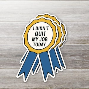 May include: A stack of stickers shaped like award ribbons. Each sticker has a yellow scalloped edge, a white center with the text "I DIDN'T QUIT MY JOB TODAY", and blue ribbons. The stickers are on a light wood grain background.
