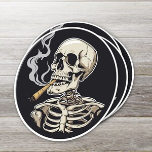May include: Oval stickers featuring a detailed illustration of a skeleton smoking a cigar. The skeleton is depicted in a relaxed pose, with a black background and white border. Smoke curls upwards from the cigar.