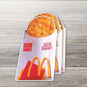 May include: Three golden-brown hash browns in white paper bags with the McDonald's logo and the words "HASH BROWNS" in red. The bags have a red and yellow design at the bottom. The background is a light wood grain.