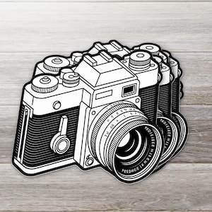 May include: Black and white sticker of a vintage camera with a lens. The camera has several dials and buttons, and the lens has a series of numbers and letters. The sticker is a graphic illustration of a classic camera.