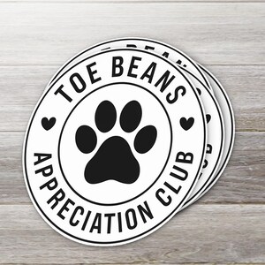 May include: Three white round stickers with black text and a black paw print. The text reads "Toe Beans Appreciation Club" with a heart on either side of the paw print.
