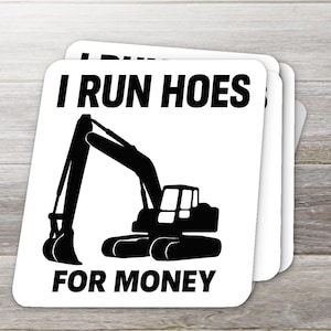 May include: A stack of white square coasters with a black excavator graphic and the text "I RUN HOES FOR MONEY". The coasters are stacked on a light brown wooden surface.