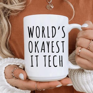 May include: White ceramic mug with the text "WORLD'S OKAYEST IT TECH" in black. The mug has a curved handle and is held by a person wearing rings. The mug is a standard size for coffee or tea.