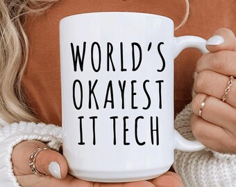 Puede incluir: Taza de cerámica blanca con el texto negro "WORLD'S OKAYEST IT TECH". La taza tiene un asa curva y la sostiene una persona con anillos. La taza es de tamaño estándar para café o té.