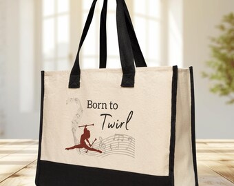 May include: Cream-colored tote bag with black trim and handles. The bag features the text "Born to Twirl" with a graphic of a baton twirler and musical notes. The bag is made of a sturdy canvas material.