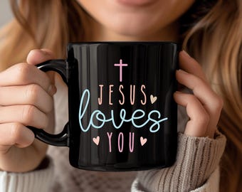 Op de afbeelding: Zwarte keramische mok met de tekst "JESUS loves YOU" in een scriptlettertype, met een roze kruis en hartjes. De mok heeft een gebogen handvat en wordt door een persoon vastgehouden.