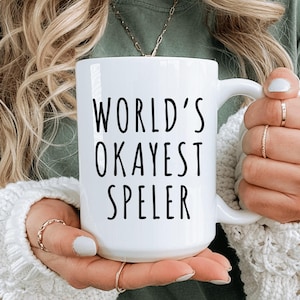 May include: White ceramic mug with the text "WORLD'S OKAYEST SPELER" in black, held by a person. The mug has a handle and is surrounded by a soft, white sweater. The person is wearing rings on their fingers.