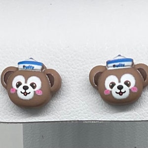 May include: A pair of brown bear-shaped stud earrings with a blue and white sailor hat. The bear has pink cheeks and a white smile.