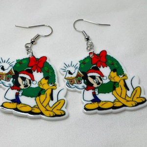 May include: A pair of white dangle earrings featuring a Christmas-themed design with Mickey Mouse and Pluto. Mickey is holding a gingerbread house and Pluto is standing next to him. They are both in front of a green wreath with a red bow.