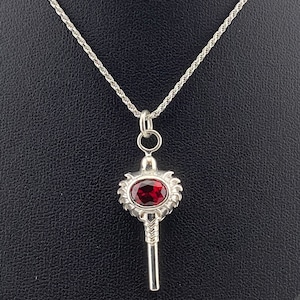 May include: A silver key pendant necklace with a red gemstone set in the key head. The chain is a silver rope chain.