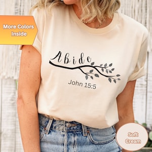 May include: A soft cream colored t-shirt with the word "Abide" in black script on a branch with gray leaves. The text "John 15:5" is printed below the branch.