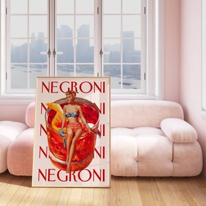 Peut inclure: Une affiche encadr&eacute;e avec une illustration de style vintage d'un cocktail Negroni. L'&oelig;uvre d'art repr&eacute;sente une femme en maillot de bain assise sur un grand verre &agrave; cocktail, avec le mot "Negroni" r&eacute;p&eacute;t&eacute; verticalement. La palette de couleurs comprend le rouge, l'orange et le beige.