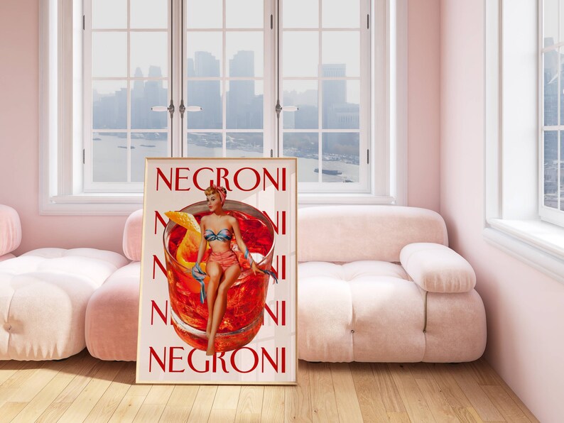 Peut inclure: Une affiche encadr&eacute;e avec une illustration de style vintage d'un cocktail Negroni. L'&oelig;uvre d'art repr&eacute;sente une femme en maillot de bain assise sur un grand verre &agrave; cocktail, avec le mot "Negroni" r&eacute;p&eacute;t&eacute; verticalement. La palette de couleurs comprend le rouge, l'orange et le beige.