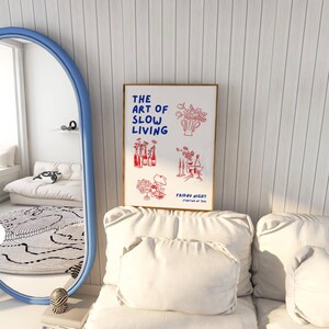 May include: A framed art print with the text "THE ART OF SLOW LIVING" in blue, and red illustrations of flowers and bottles. The print is displayed on a white couch with pillows, near a blue-framed mirror.