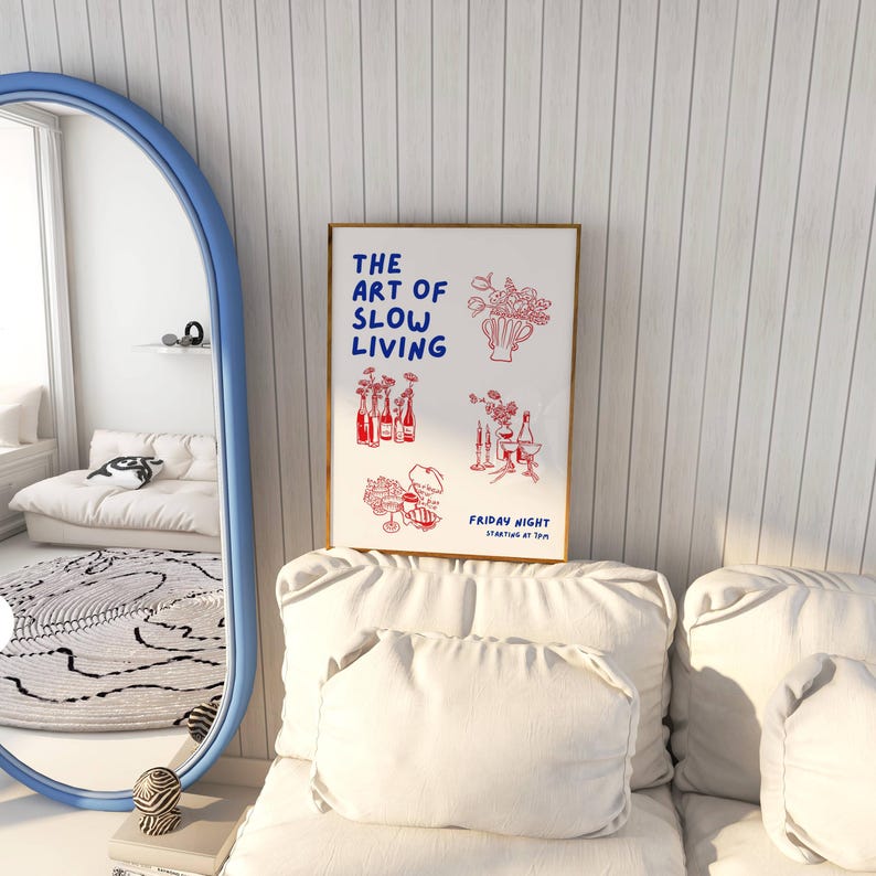 May include: A framed art print with the text "THE ART OF SLOW LIVING" in blue, and red illustrations of flowers and bottles. The print is displayed on a white couch with pillows, near a blue-framed mirror.