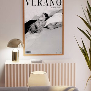 May include: A black and white photograph of a couple lying on a beach. The image is framed in a wooden frame and has the text "VERANO a vintage summer" printed on it.