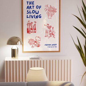 May include: A framed art print with the text "THE ART OF SLOW LIVING" in blue and red illustrations of flowers, bottles, and food. The print is displayed above a white cabinet with a gold lamp and a beige lampshade.