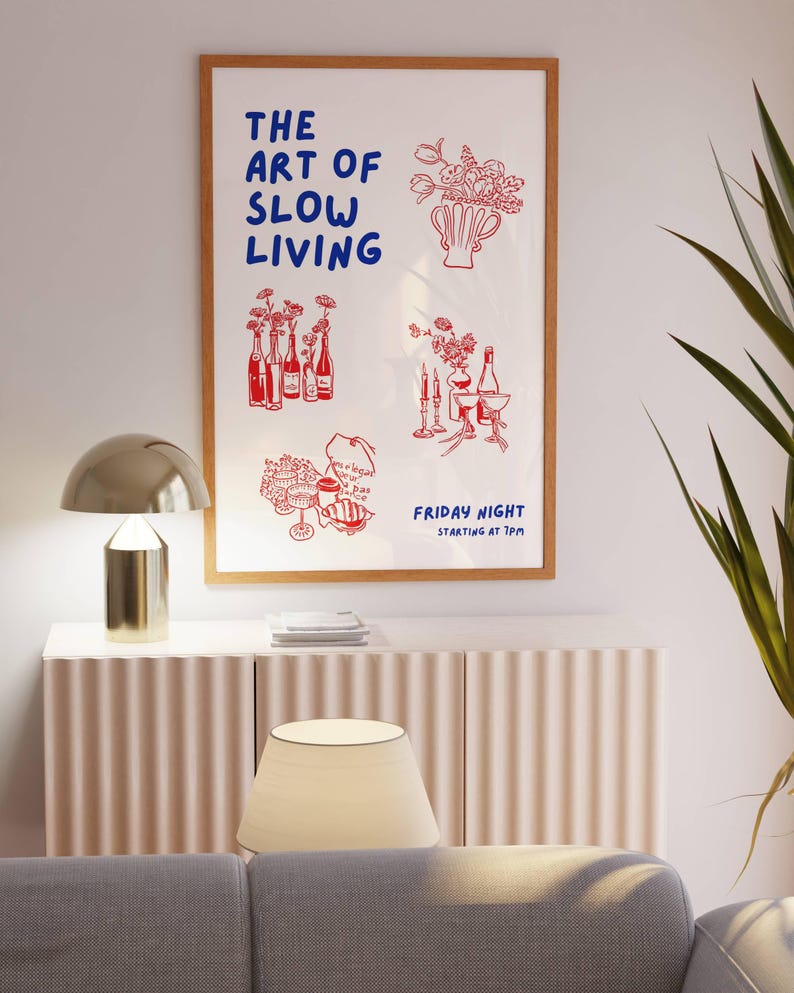 May include: A framed art print with the text "THE ART OF SLOW LIVING" in blue and red illustrations of flowers, bottles, and food. The print is displayed above a white cabinet with a gold lamp and a beige lampshade.