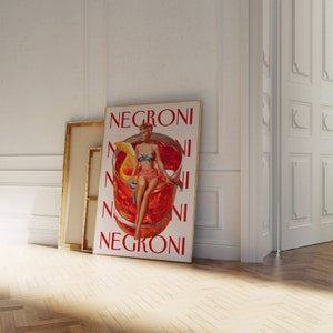 Peut inclure: Une affiche encadr&eacute;e avec le mot "NEGRONI" en rouge, repr&eacute;sentant une illustration vintage d'une femme en bikini assise dans un verre &agrave; cocktail. L'affiche est appuy&eacute;e contre un mur blanc et un sol en bois.