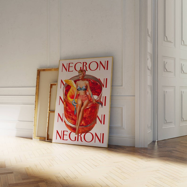 Peut inclure: Une affiche encadr&eacute;e avec le mot "NEGRONI" en rouge, repr&eacute;sentant une illustration vintage d'une femme en bikini assise dans un verre &agrave; cocktail. L'affiche est appuy&eacute;e contre un mur blanc et un sol en bois.