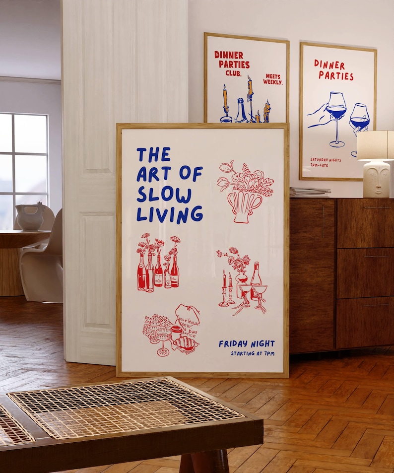 May include: A framed art print with the text "THE ART OF SLOW LIVING" in blue, and illustrations in red. Additional framed prints with "DINNER PARTIES" text and illustrations are visible. The art features images of flowers, bottles, and wine glasses.