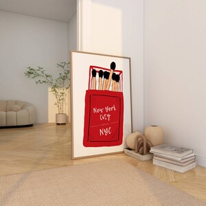 May include: A red matchbox with the text "New York City Classic NYC" printed on the front. The matchbox is filled with matches with black heads.