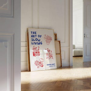 May include: A framed poster with the text "THE ART OF SLOW LIVING" in blue, with red illustrations of flowers, bottles, and glasses. The poster is leaning against a wall in a room with a wooden floor.