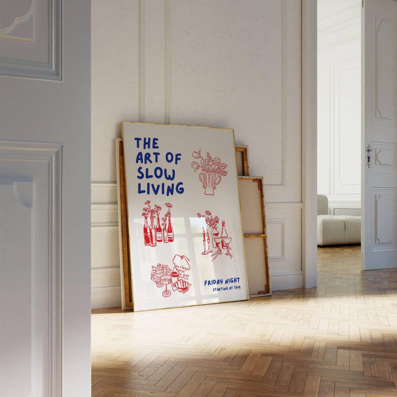 May include: A framed poster with the text "THE ART OF SLOW LIVING" in blue, with red illustrations of flowers, bottles, and glasses. The poster is leaning against a wall in a room with a wooden floor.