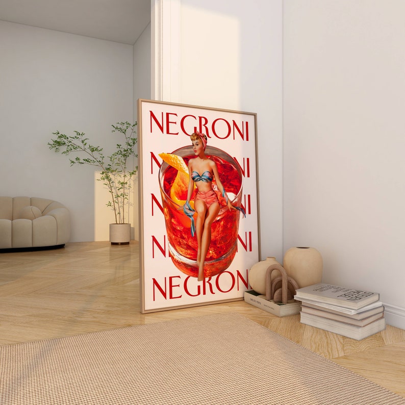 Peut inclure: Une affiche encadr&eacute;e avec le mot "NEGRONI" en rouge. L'affiche pr&eacute;sente une illustration vintage d'une femme en maillot de bain assise dans un verre &agrave; cocktail avec une tranche d'orange. L'affiche est appuy&eacute;e contre un mur blanc.