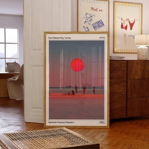 May include: Wooden framed art prints featuring beach scenes and retro illustrations. One print shows a large red sun over a beach with figures. Other prints display beach-themed drawings with text. The prints are displayed in a room with wooden floors.
