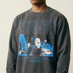 Puede incluir: Sudadera gris oscuro con la palabra "HEAT" en azul y un gráfico de una figura enmascarada. La sudadera también presenta una gorra de béisbol beige.