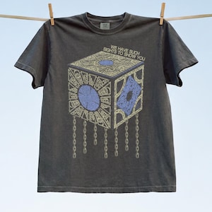 Könnte beinhalten: Ein anthrazitfarbenes Kurzarm-T-Shirt mit einer Grafik einer goldenen Puzzle-Box mit blauen kreisförmigen und rautenförmigen Akzenten. Der Text "WE HAVE SUCH SIGHTS TO SHOW YOU" ist über der Box gedruckt, die mit hängenden Metallketten versehen ist.