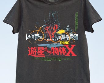 Può includere: Una t-shirt grigio antracite a maniche corte con una stampa grafica del film The Thing. Il design include una fila di ritratti di personaggi, testo giallo e rosso e caratteri giapponesi. La maglietta è appesa a un filo per il bucato.